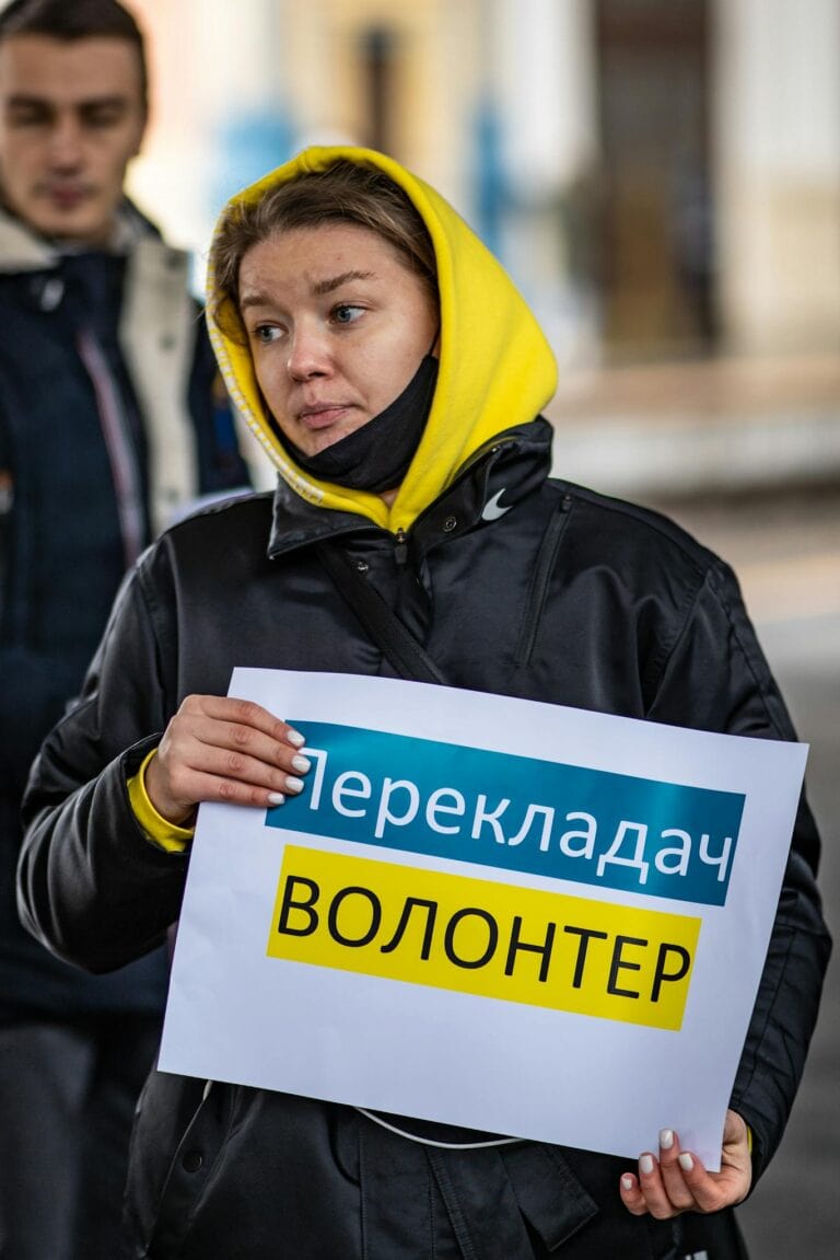 A woman in a yellow hoodie holding a volunteer translation sign outdoors in Budapest.